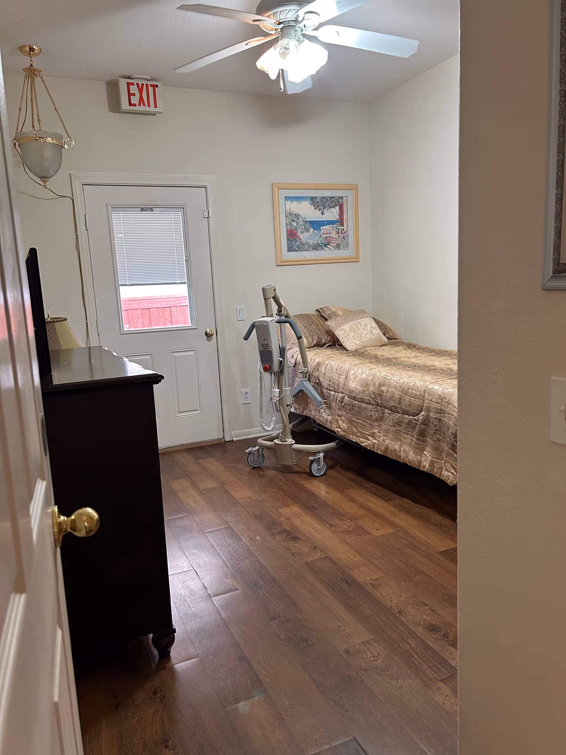 A small bedroom in a senior facility with a bed, walker, dresser, and exit door.