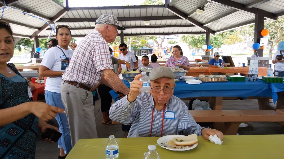 Elderly man gives thumbs up at an outdoor senior event under a pavilion with volunteers.