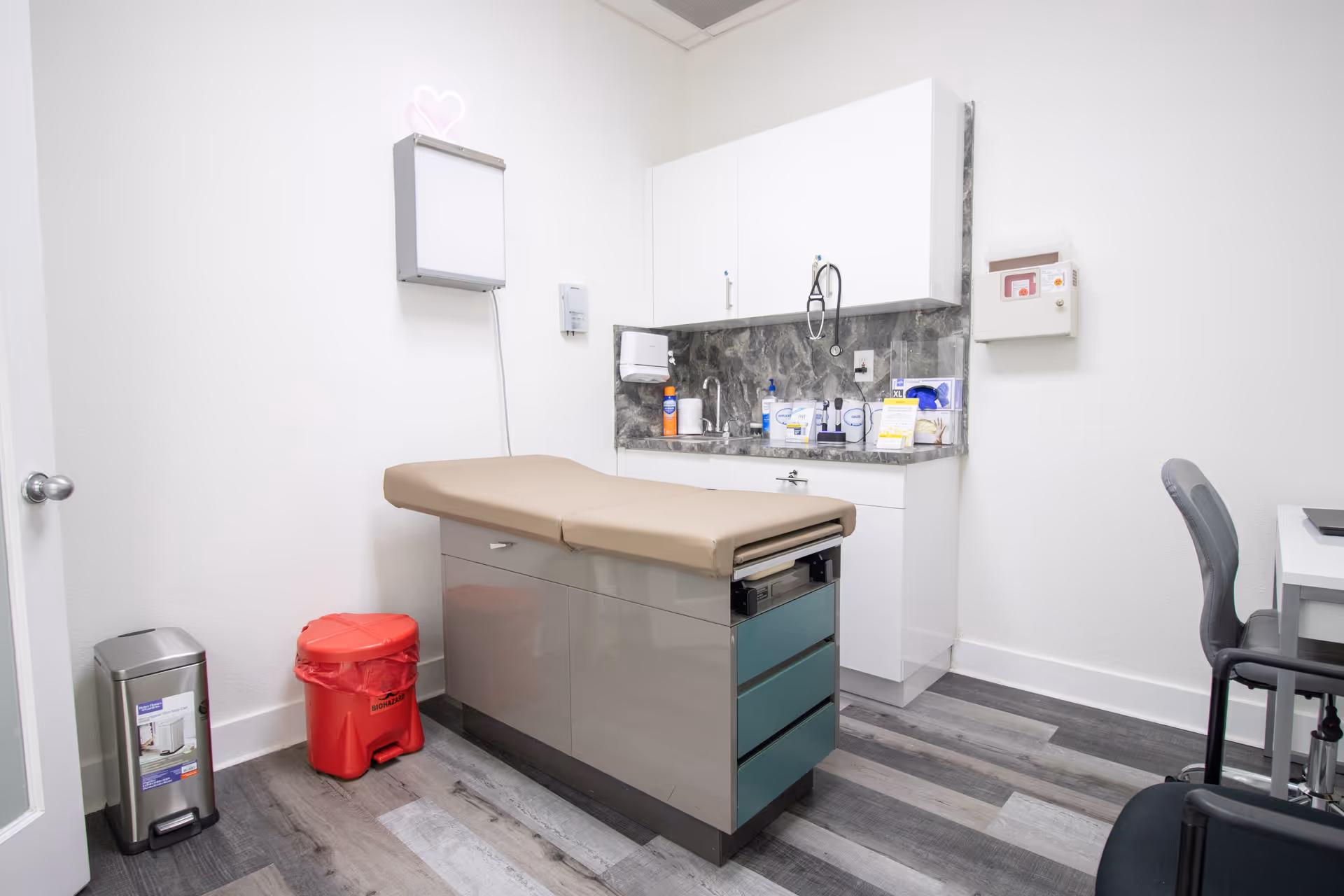 Clinical examination room showing a beige medical table, cabinets, sink, and chairs.