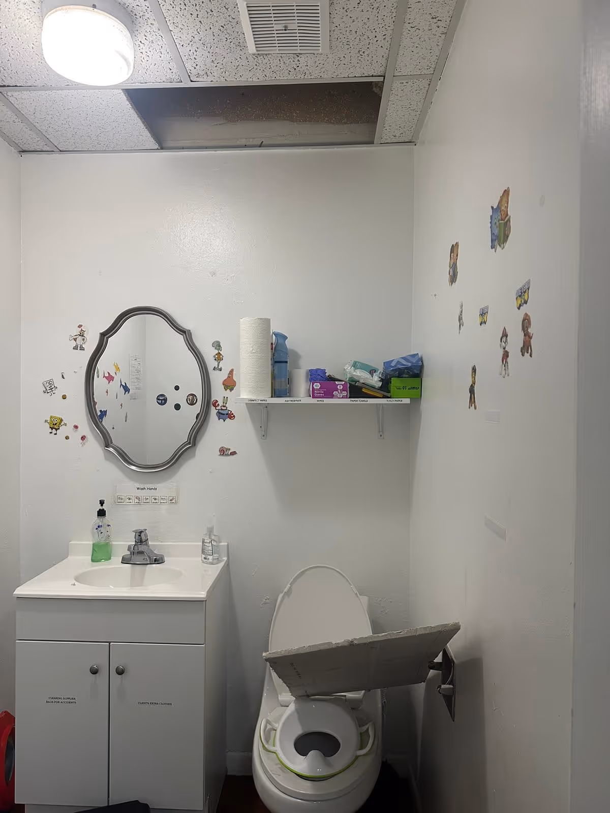 Small bathroom with sink, mirror, toilet, shelf of supplies, wall decals, and a missing ceiling panel.