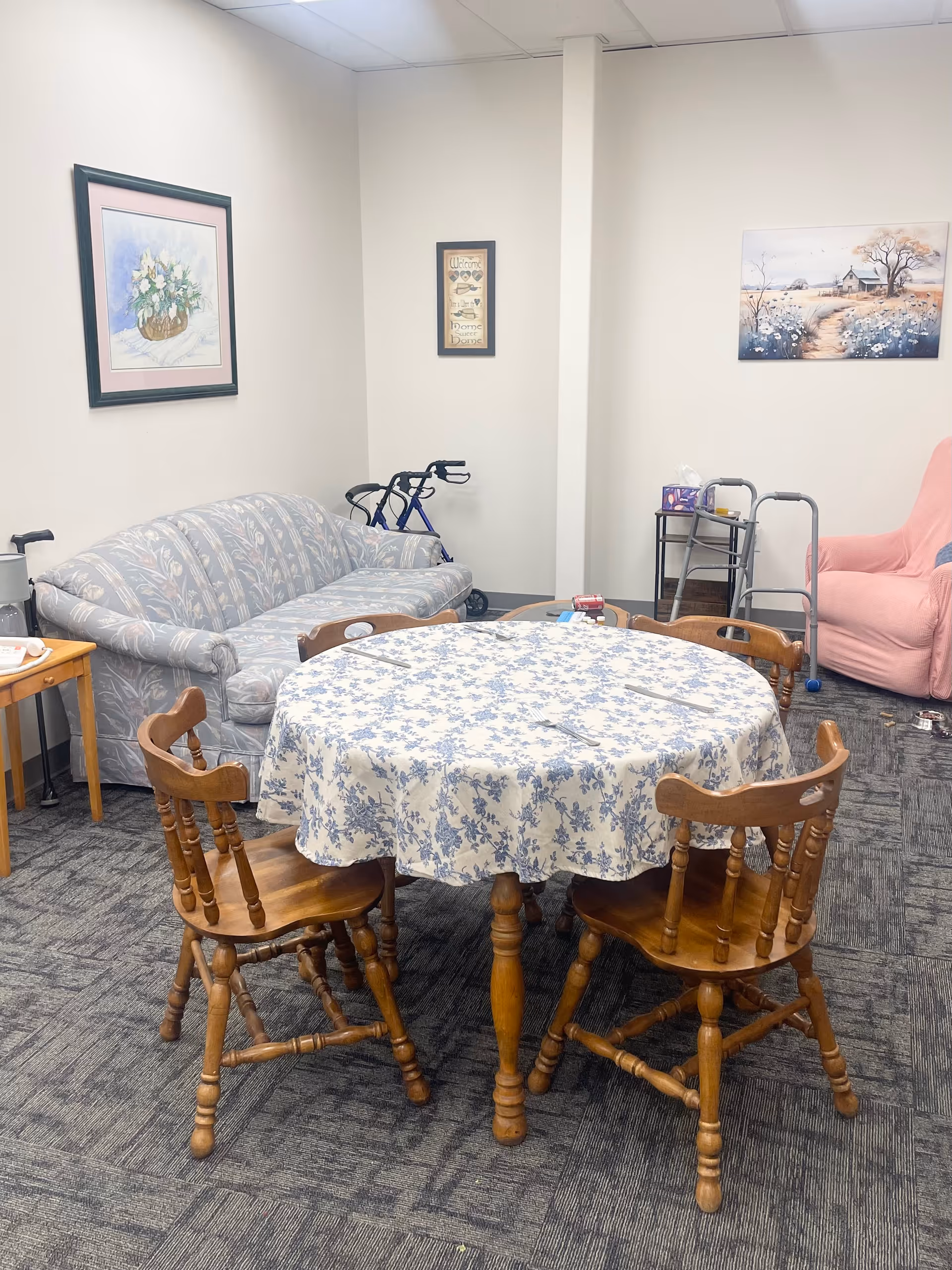 Cozy lounge area with a round floral table, wooden chairs, sofa, and walkers nearby.