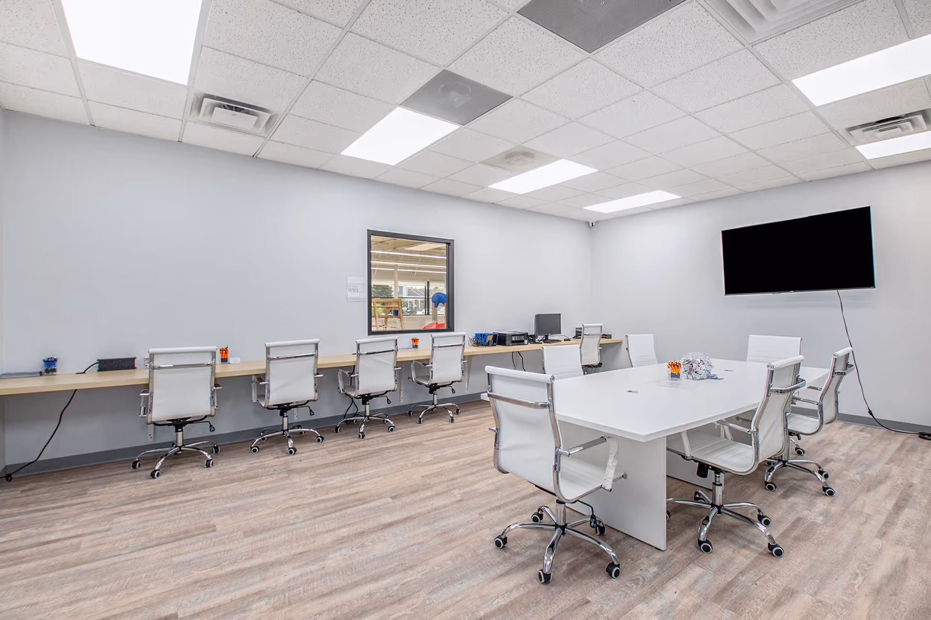 Bright interior room with a large white table, white chairs, and a wall-mounted TV.
