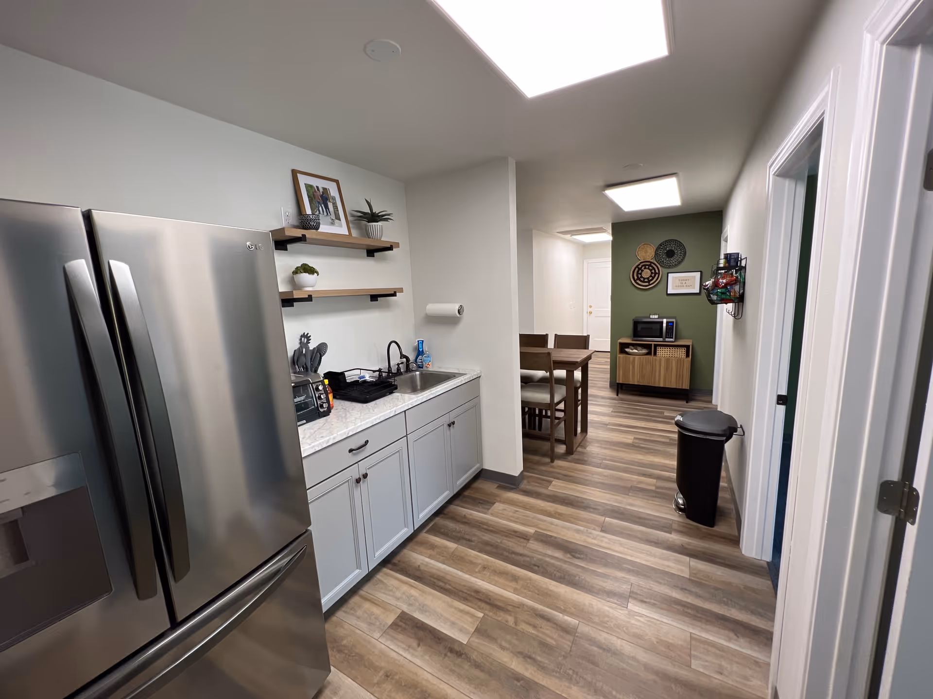 Modern kitchen area in a senior living facility with stainless fridge, sink, shelves, and dining space.