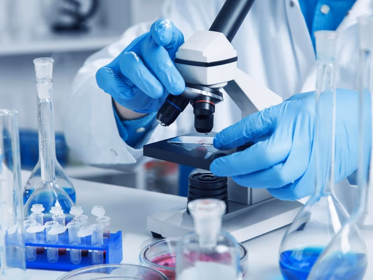 Gloved hands operate a microscope beside glassware and vials in a laboratory.
