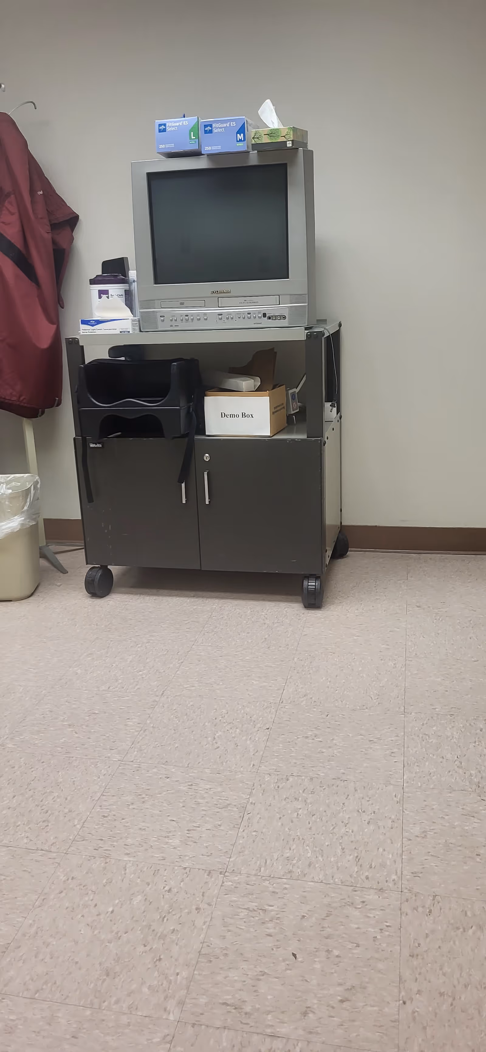 A medical cart with an old TV sits against a plain hallway wall, tissue boxes atop.