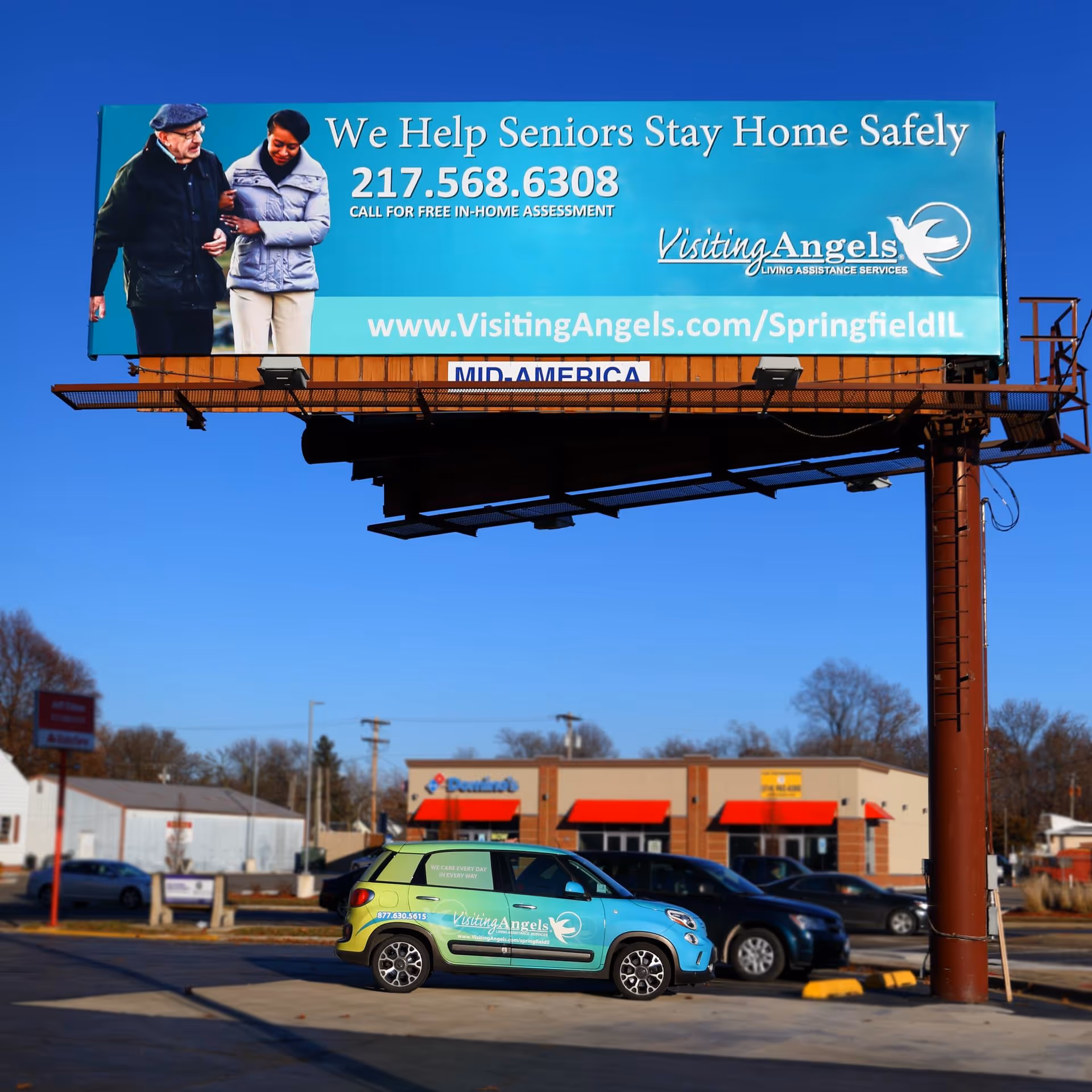 A large Visiting Angels billboard overlooks a parking lot with a teal branded car.