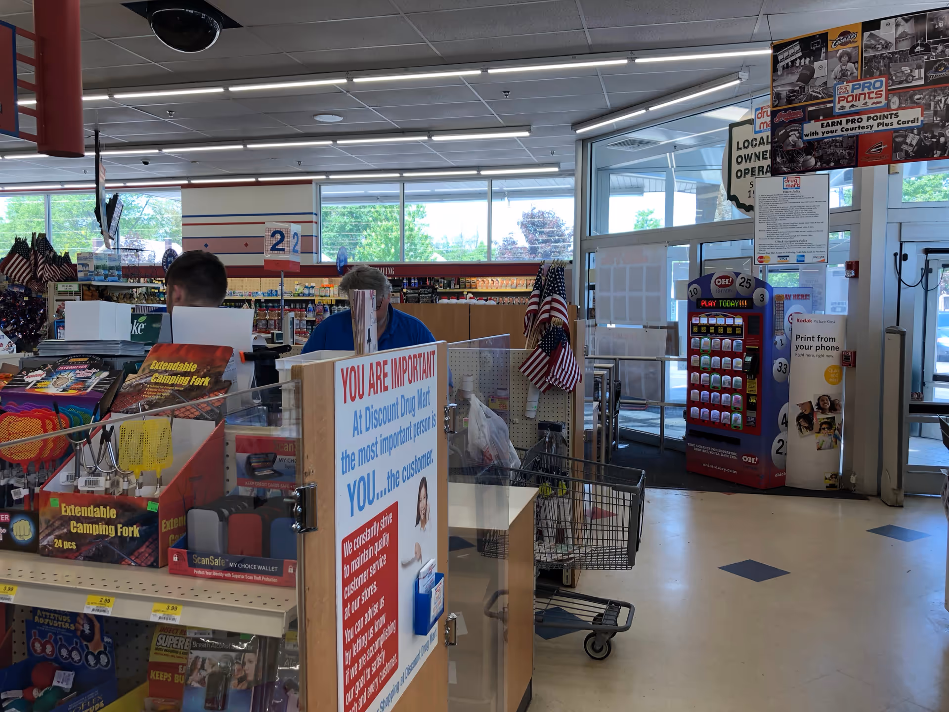 Inside Discount Drug Mart checkout area with two clerks and shopping carts near a sign.