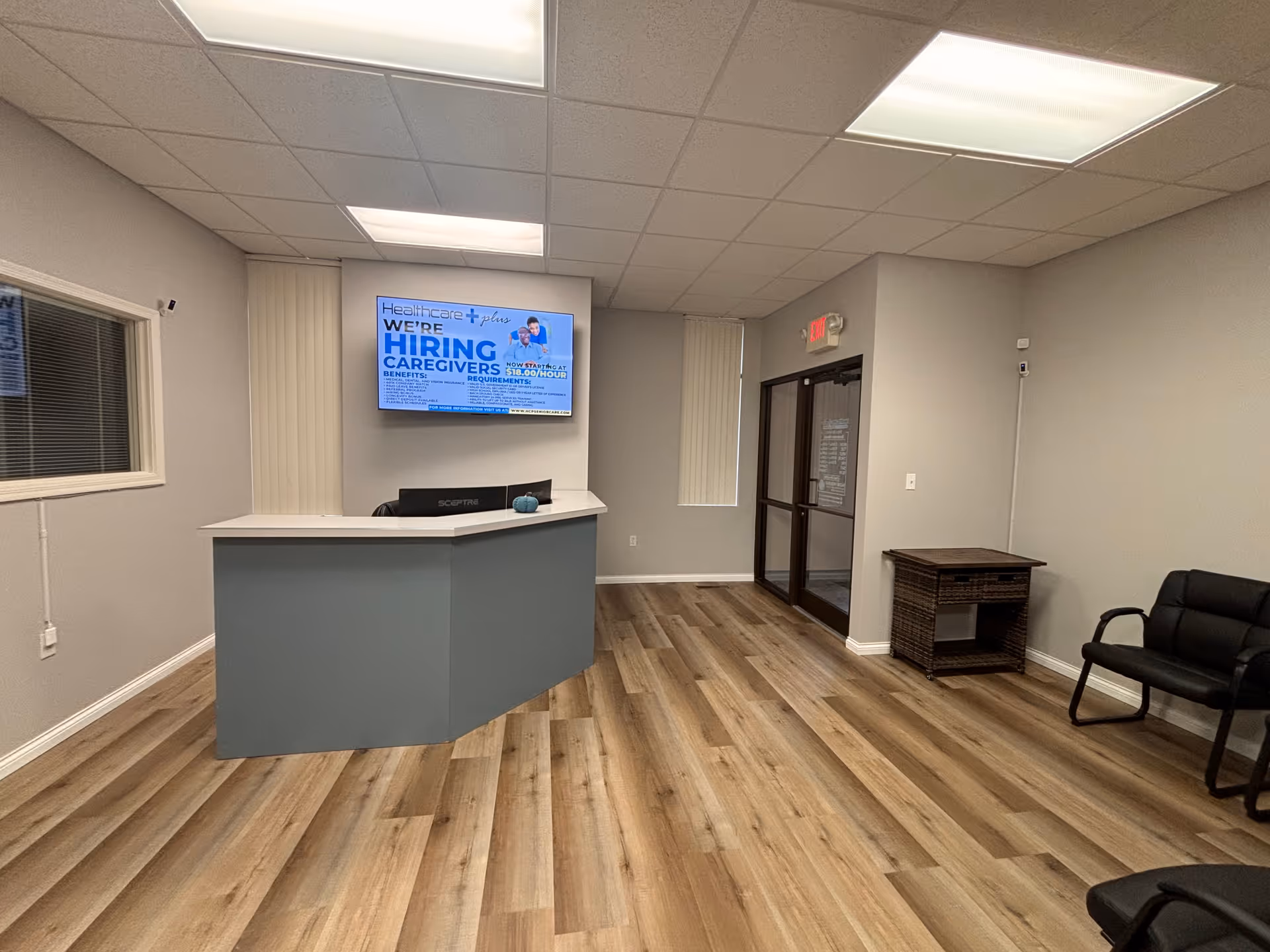 Reception desk with a screen, seating area, and doorway in a modern senior living lobby.
