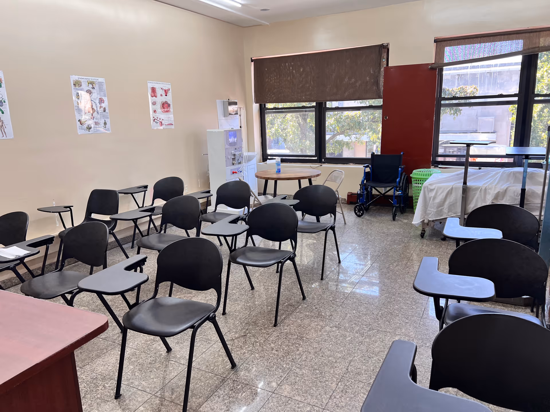 Empty classroom-style room with rows of black chairs with attached desks, posters, windows, and wheelchair.