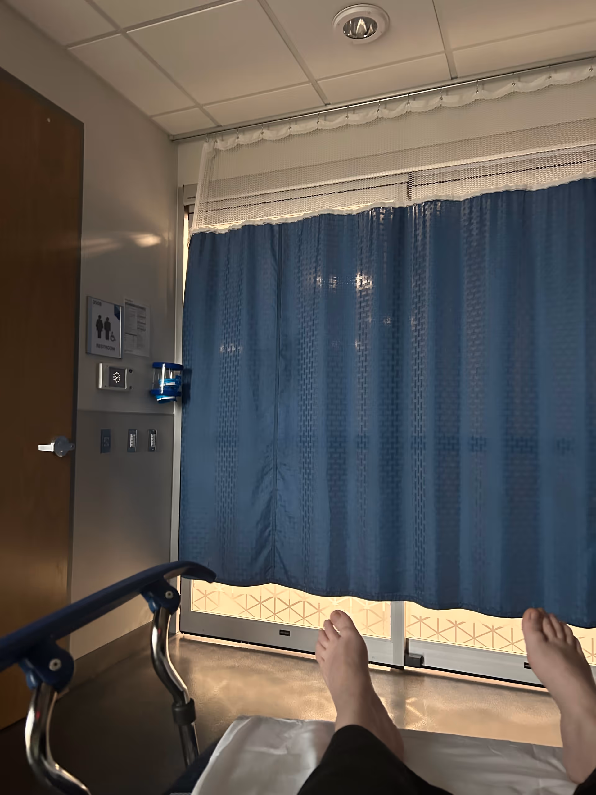 Hospital room with blue privacy curtain drawn around bed; patient legs visible at foot.