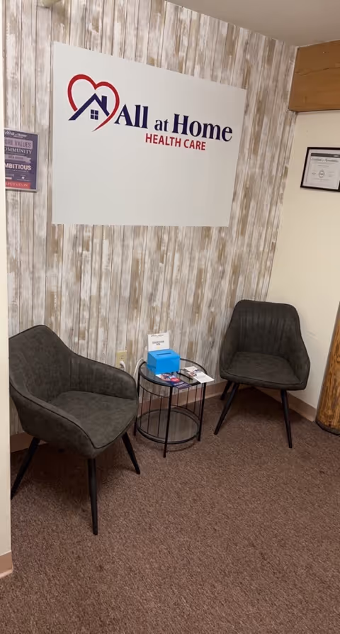 Waiting area with two gray chairs and a table beside an 'All at Home Healthcare' sign.