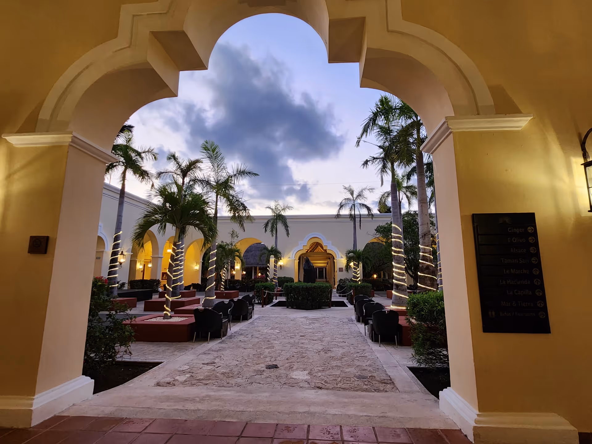 Arcaded outdoor courtyard with arches, palm trees, and string lights.