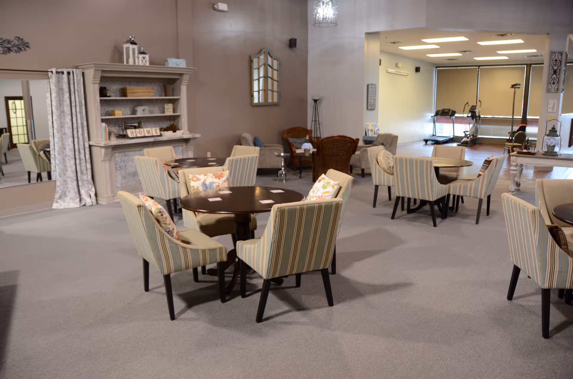Spacious communal lounge with striped chairs around round tables and exercise area visible in back.