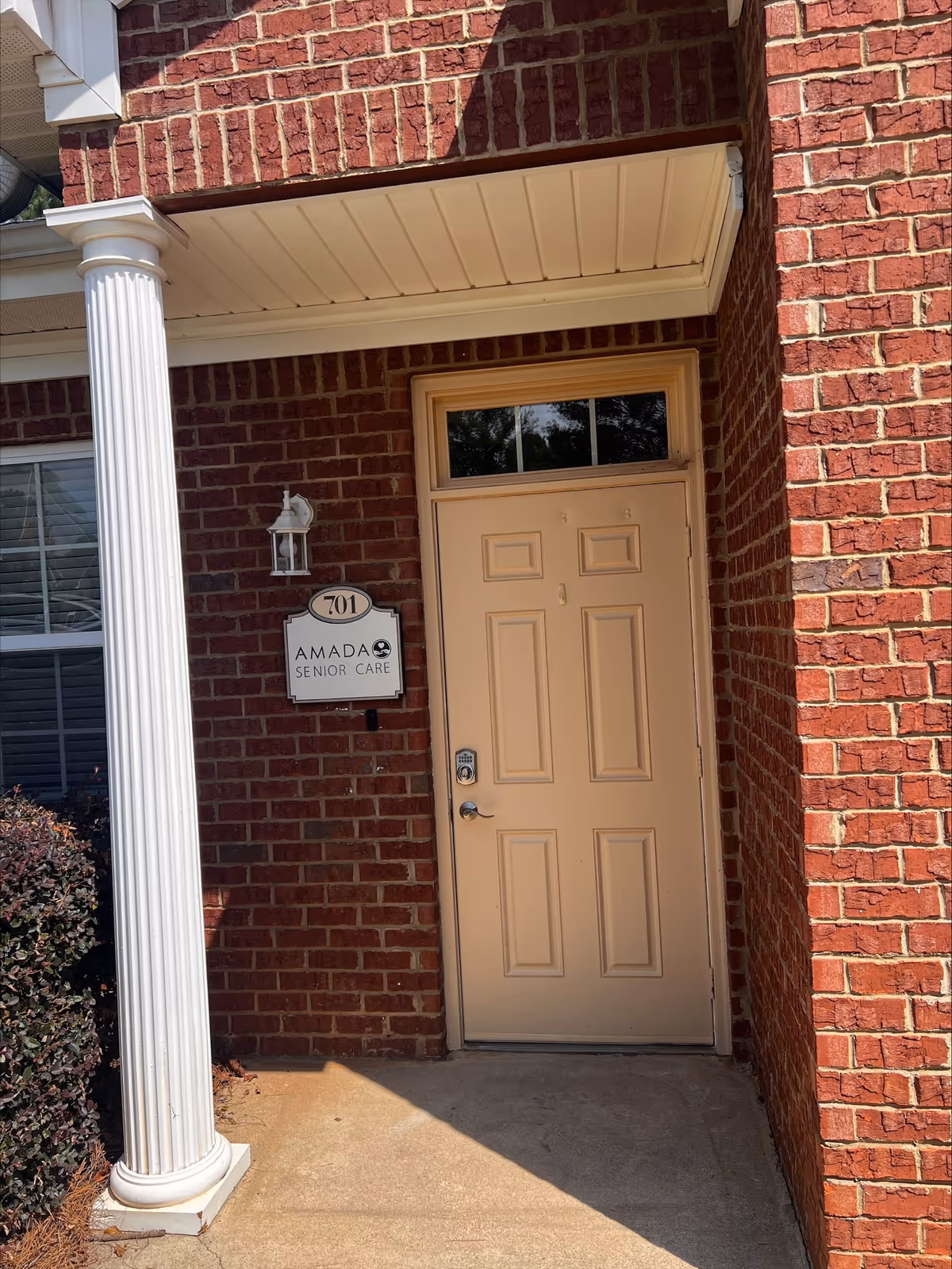 Brick building entrance with white column, beige door, and Amada Senior Care sign.