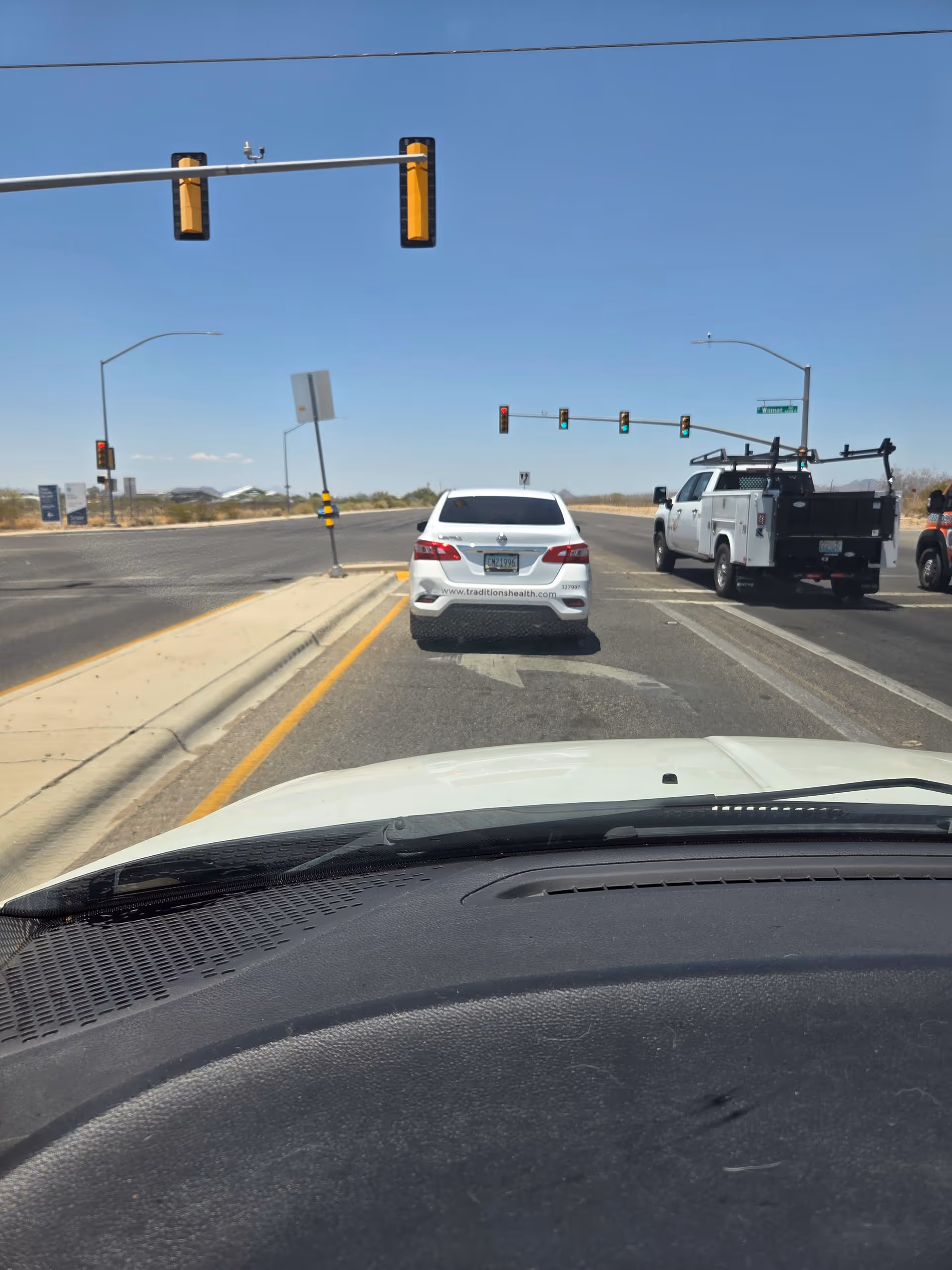 View from inside a car at an intersection, white car ahead bearing Traditions Health logo.