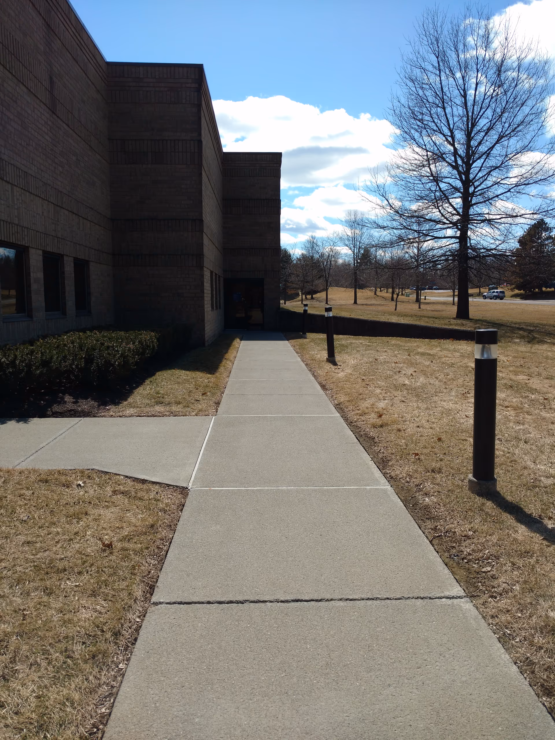 Concrete sidewalk leads to a brick senior living building entrance with leafless trees.