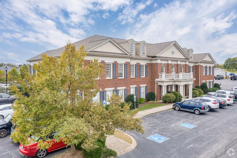 Brick senior living building with a covered entrance, parking, and trees in foreground.