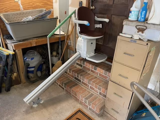 Stair lift chair installed on brick steps in a cluttered indoor utility area.