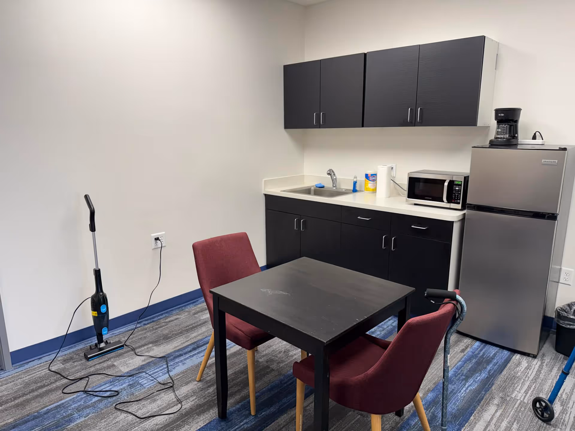 Small facility kitchenette with dark cabinets, table, chairs, microwave, fridge, and vacuum cleaner.