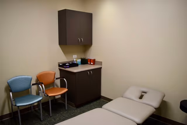 Therapy room with a treatment bed, two chairs, and dark cabinets along beige walls.