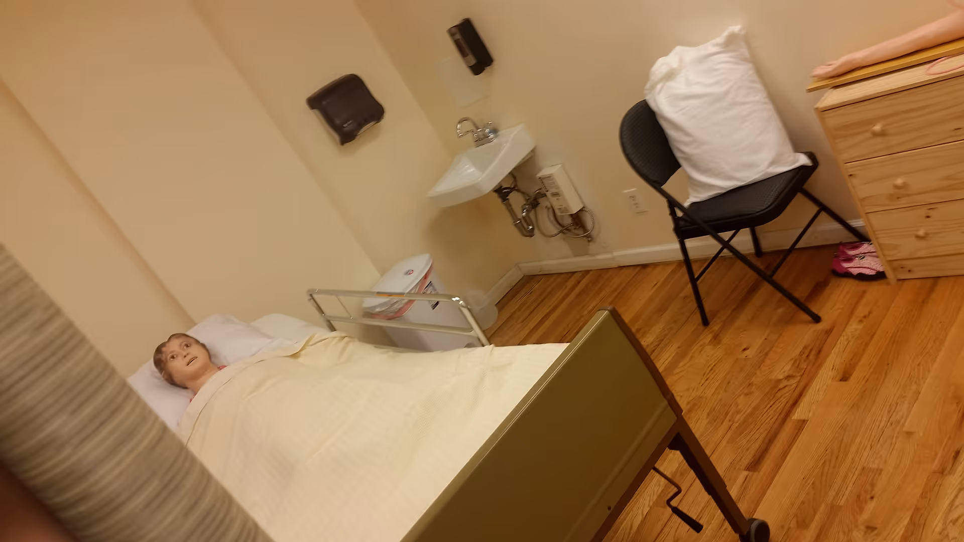 Resident rests in a small bedroom-like care room with bed, chair, and sink.