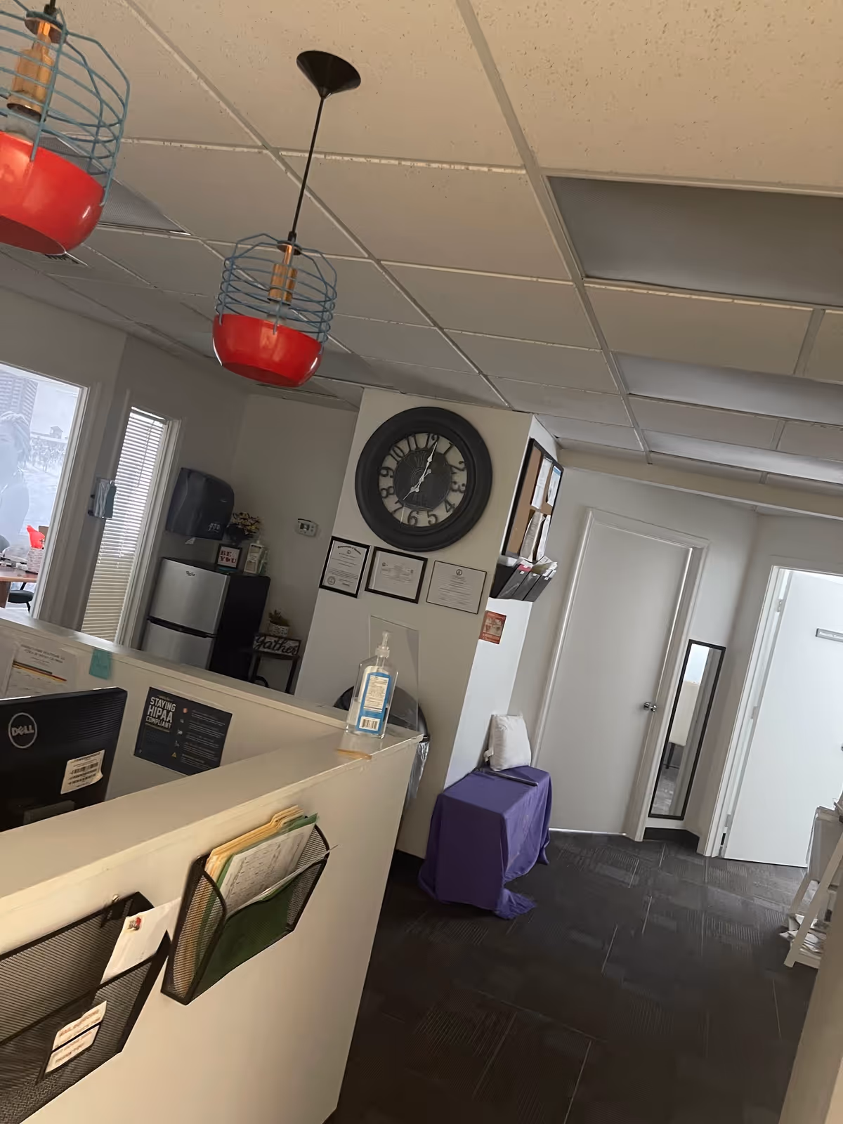 Reception area with a clock, certificates, desk, sanitizer, and purple-covered furniture.