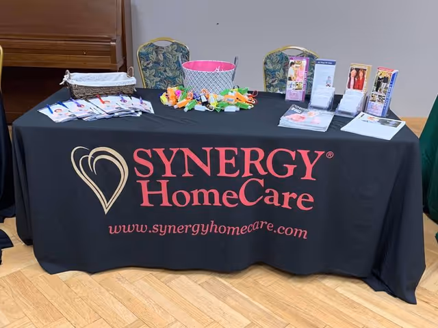 A Synergy HomeCare promotional table indoors with brochures and colorful items on a black tablecloth.