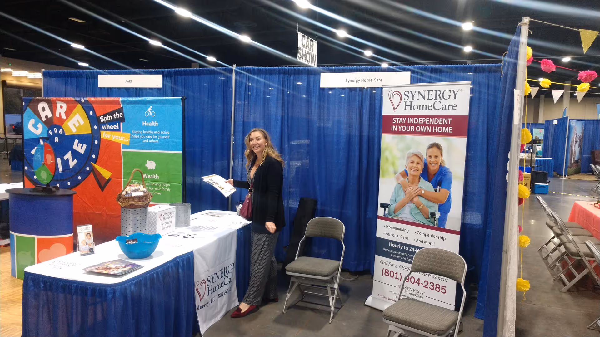 Indoor expo booth for SYNERGY HomeCare of Alta with staff member, table, and banners.