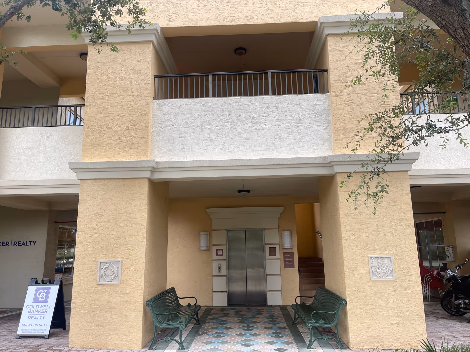 Beige building entrance with elevator, columns, and green benches outdoors.