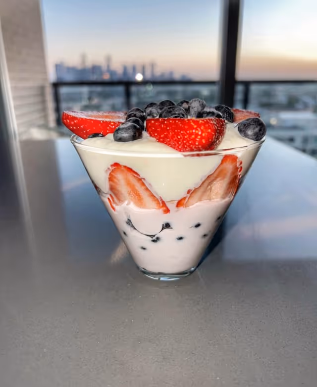 A glass dessert topped with strawberries and blueberries on a balcony at sunset.
