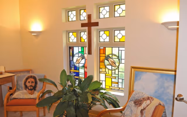 Small interior chapel-like sitting area with stained glass, a cross, chairs, and plants.