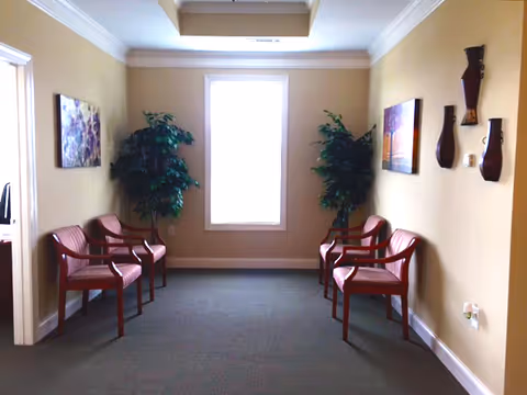 A beige-walled waiting area corridor with six red chairs, plants, and a window.
