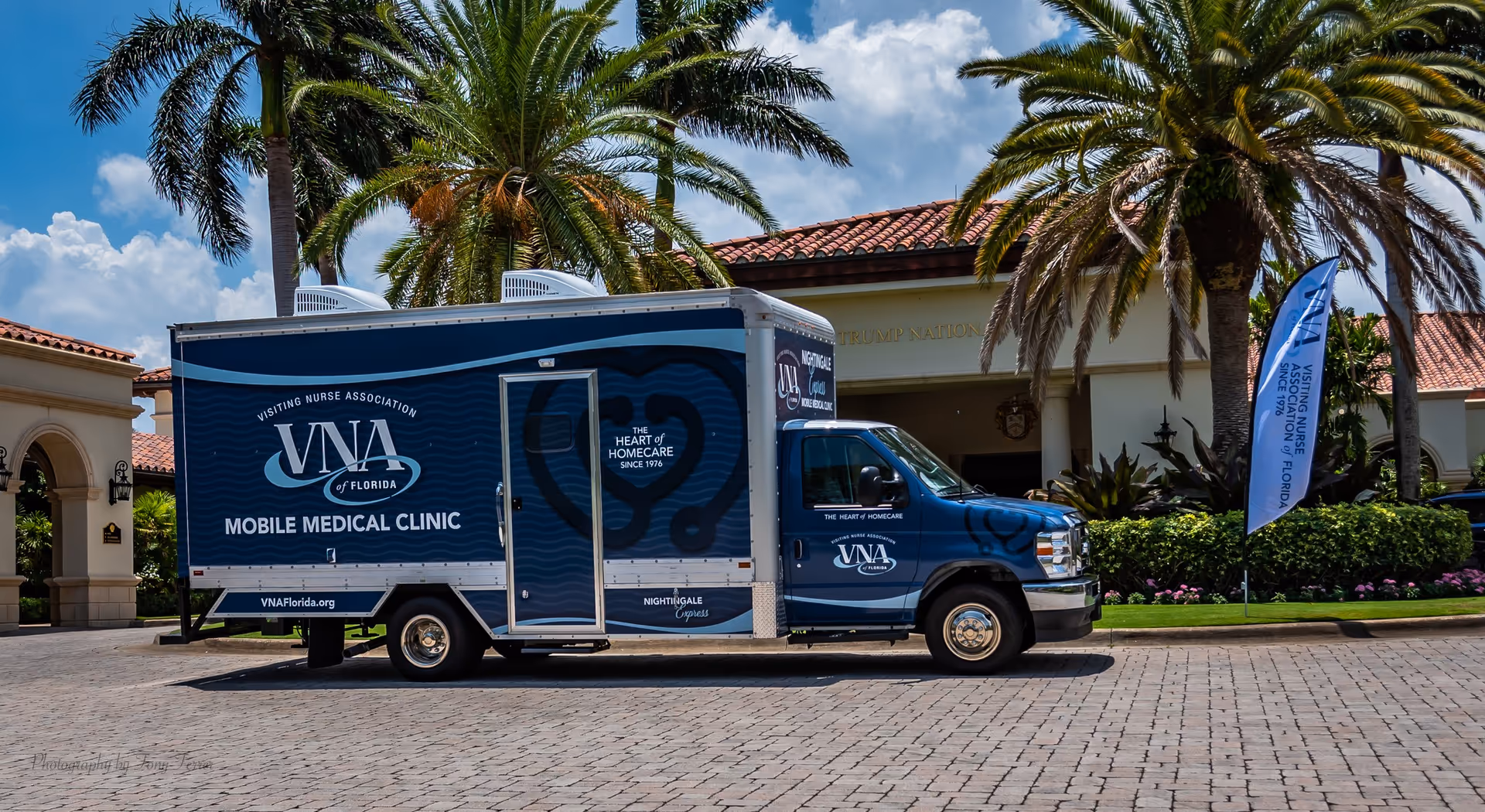 Blue Visiting Nurse Association mobile clinic van parked outside a tropical building with palms.