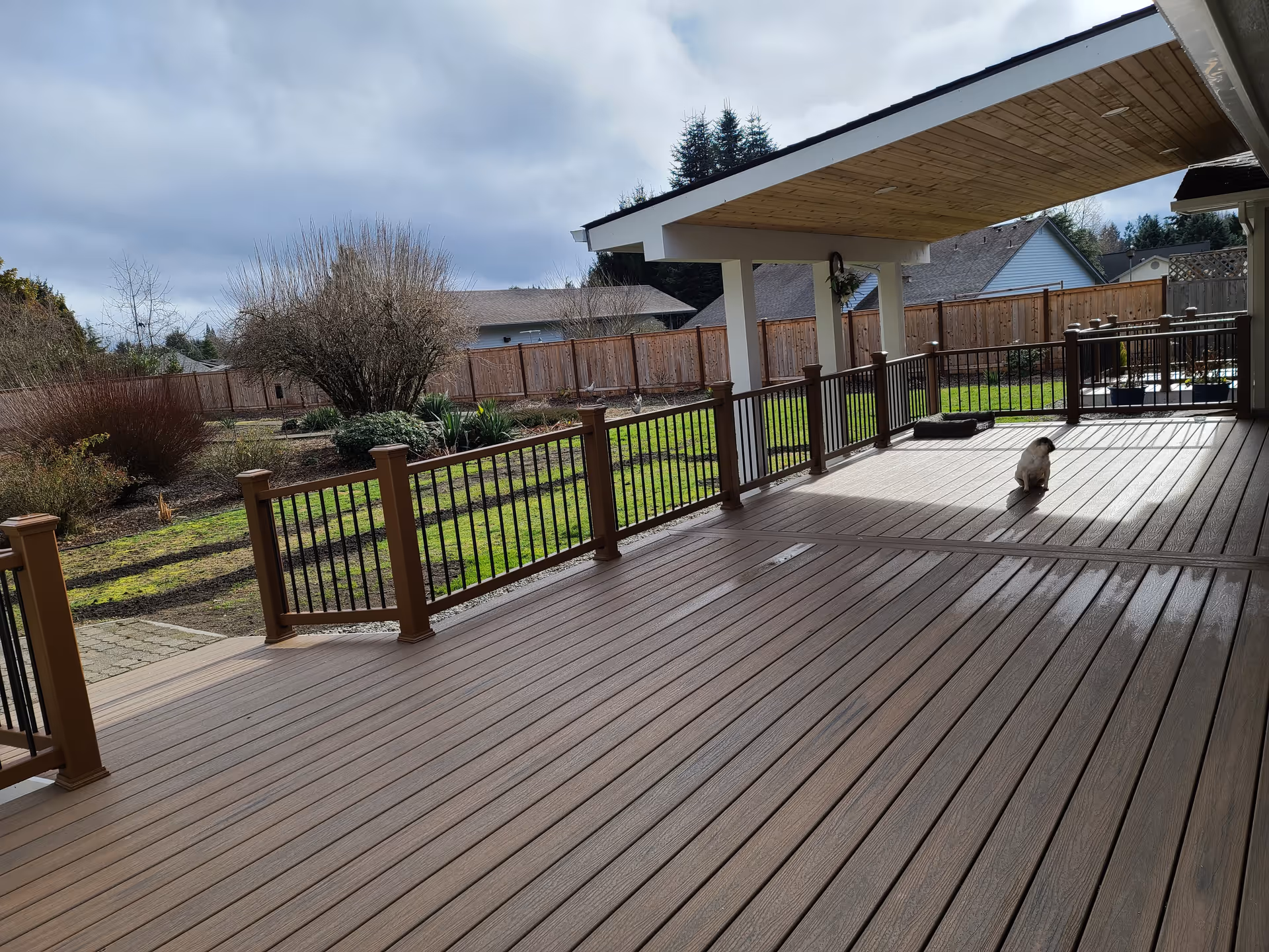 Wooden deck with brown railing, fenced yard beyond, and a small dog near the house.