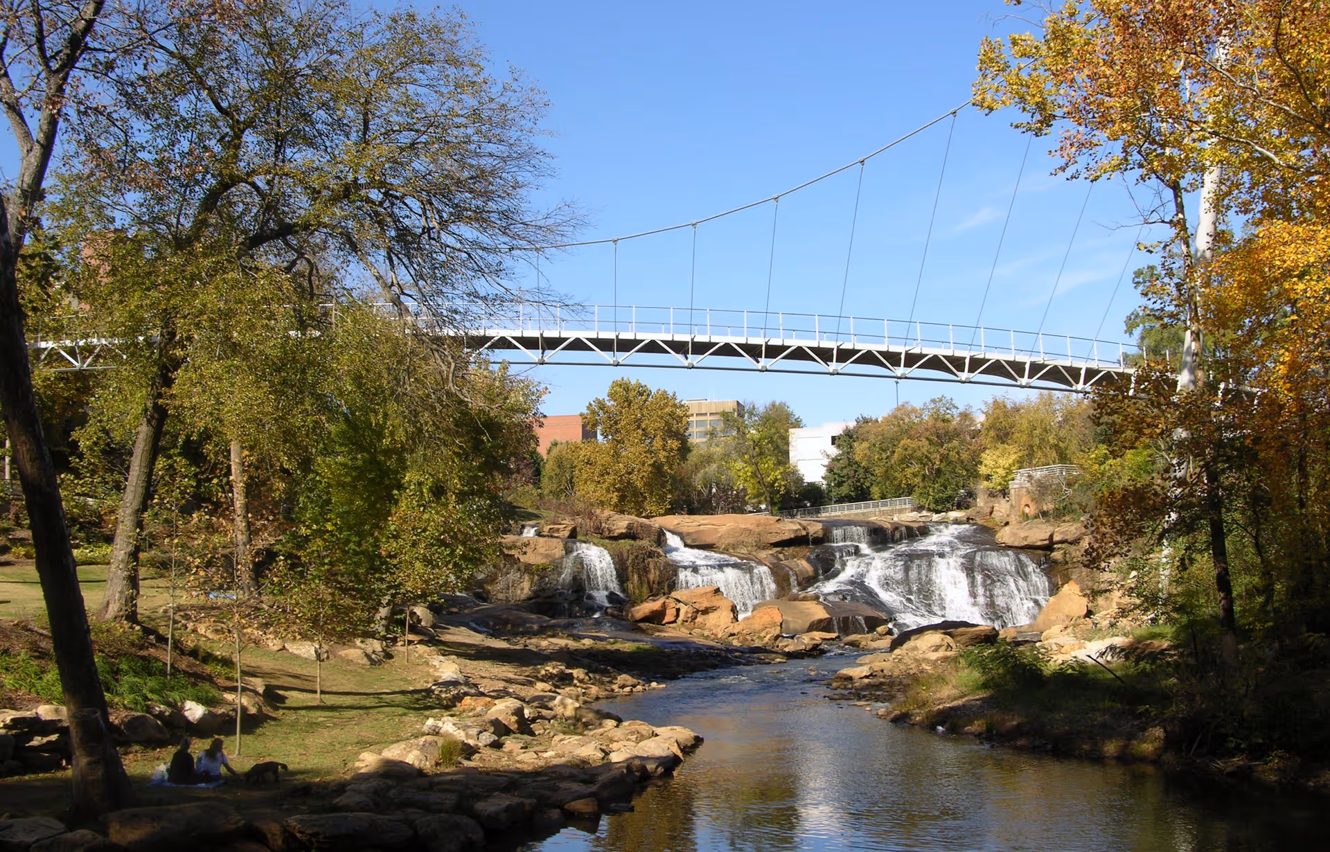 Suspension bridge spans a river with waterfall, surrounded by autumn-colored trees.