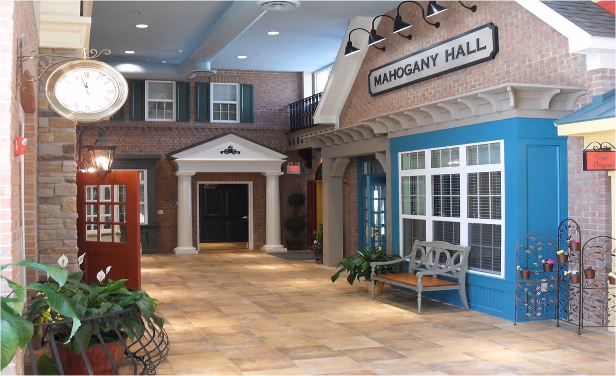 Indoor shopping-style corridor with brick walls, storefronts, benches, and potted plants.