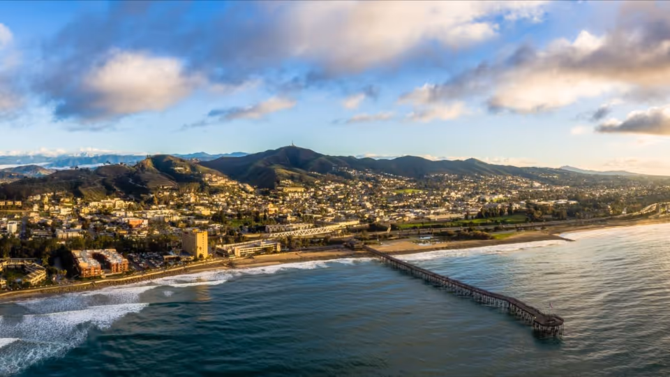 Aerial view of a coastal town with hills and a long pier extending into the ocean