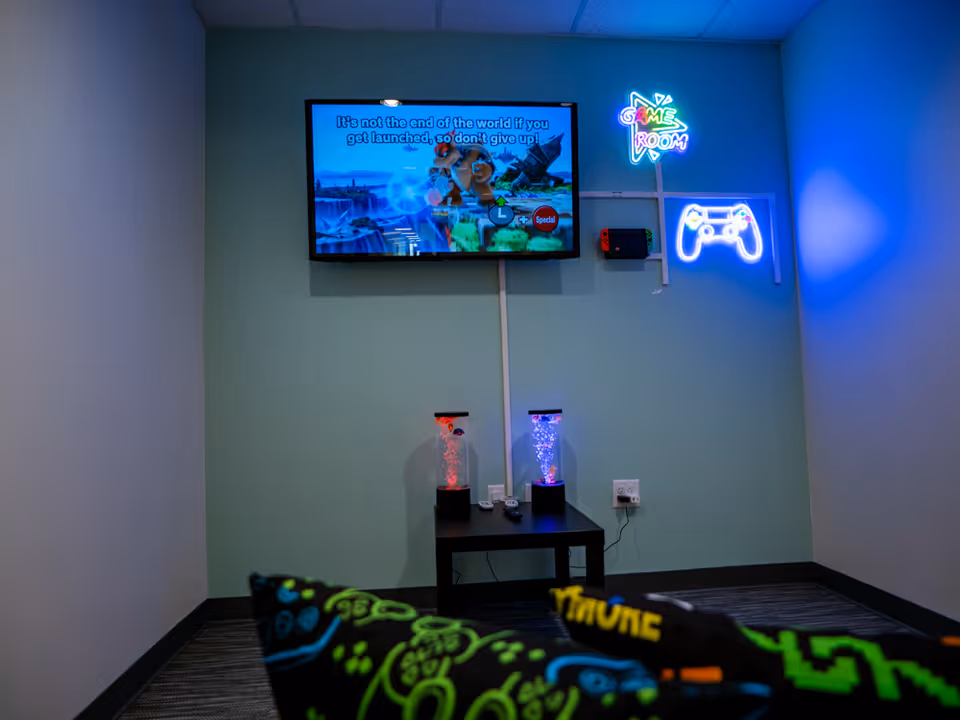 A dimly lit game room with a wall-mounted TV, neon signs, lava lamps, and colorful blankets.