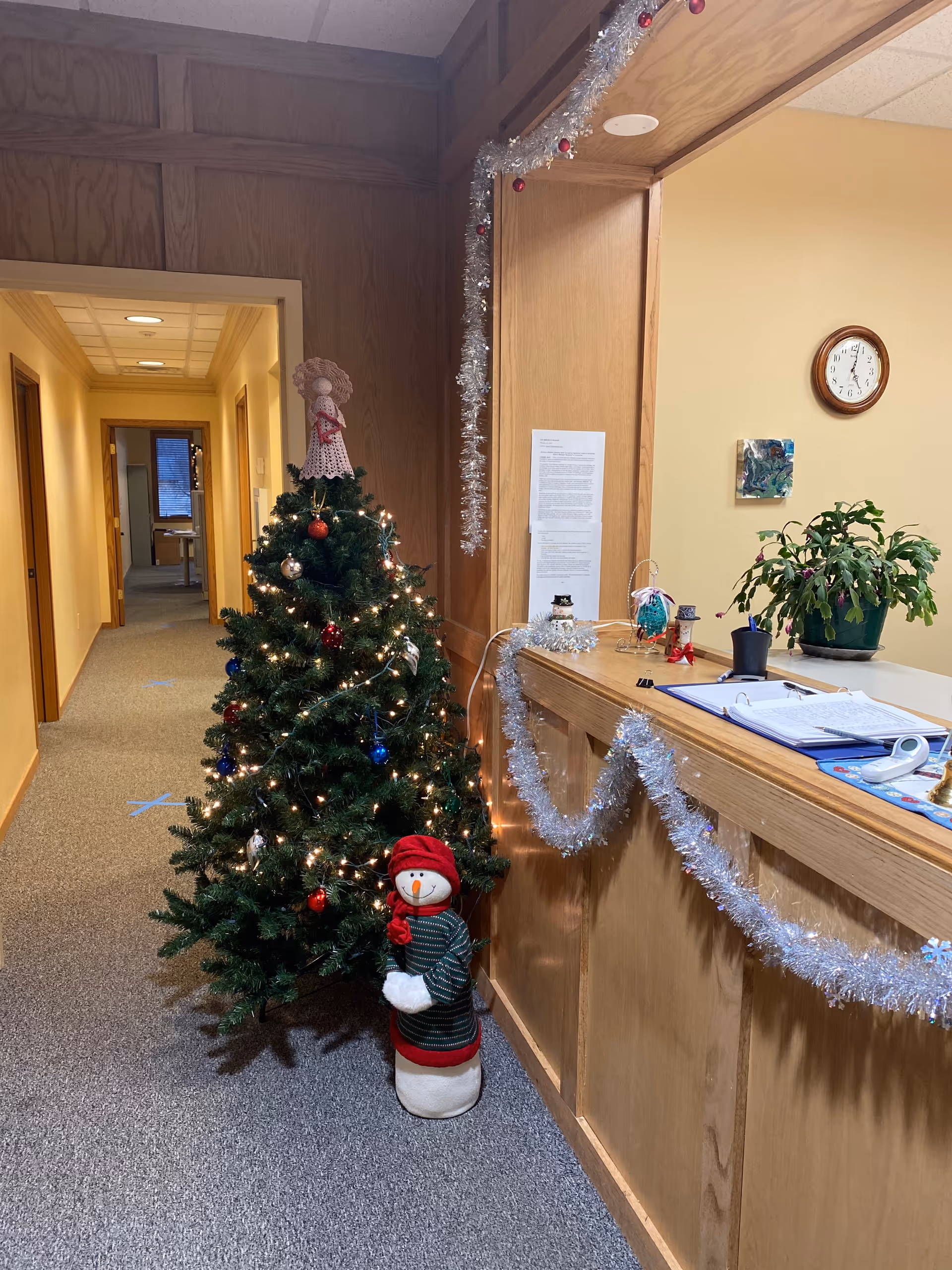 Holiday-decorated interior hallway with a Christmas tree, snowman figure, and garland near a reception desk.