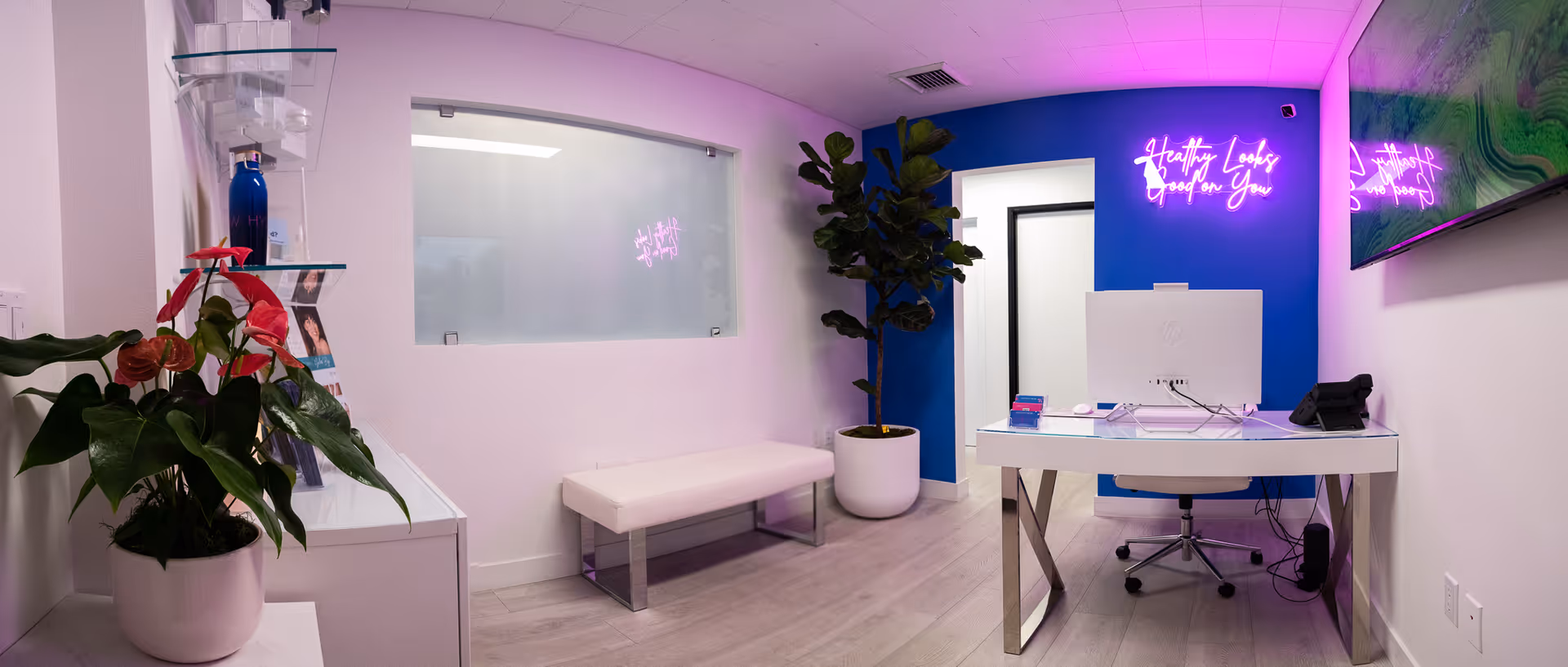 Modern interior featuring a white reception desk, neon sign, bench, and plants in a hydration clinic.