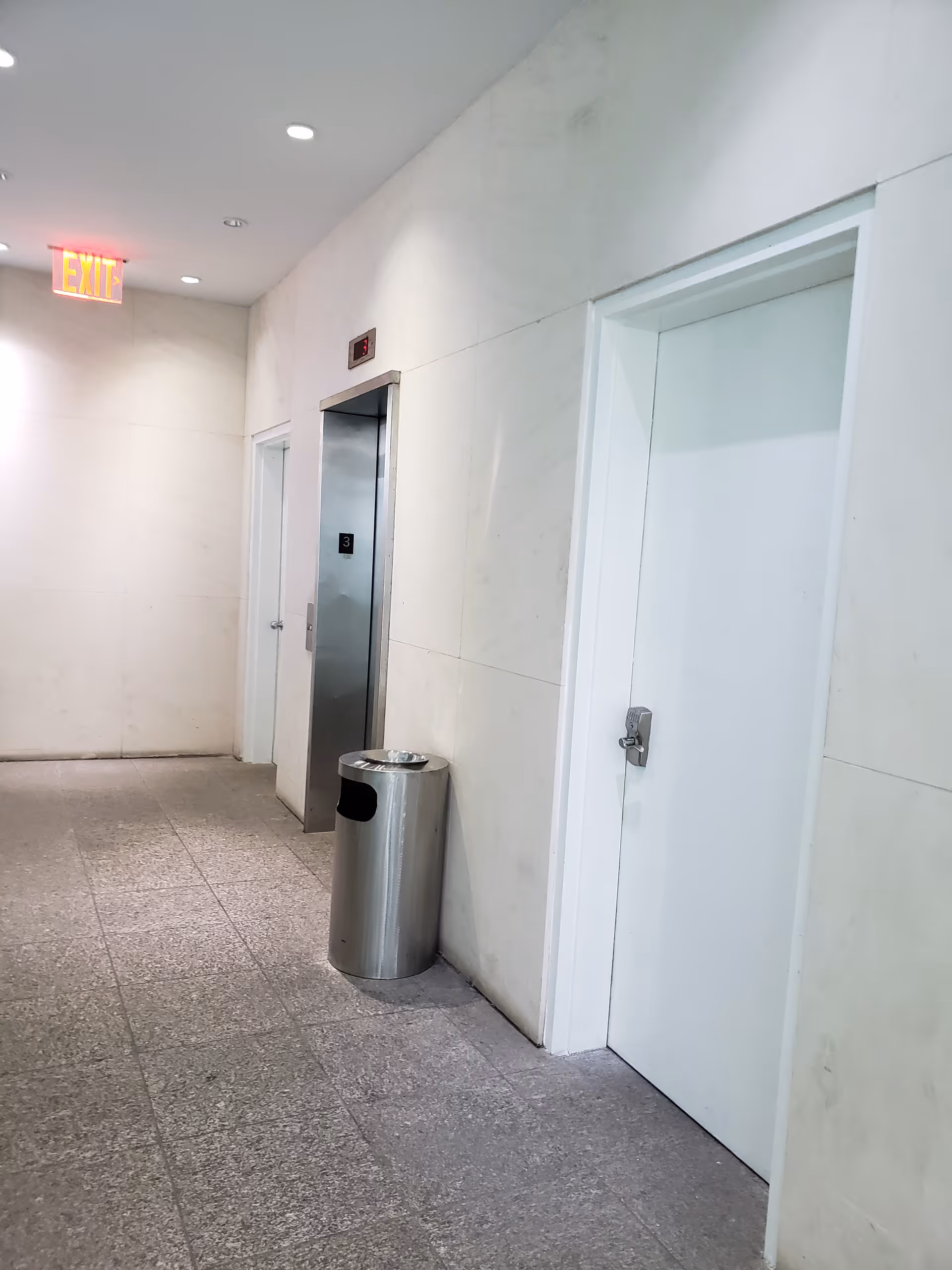 Marble-walled hallway with an elevator, exit sign, and a metal trash can.