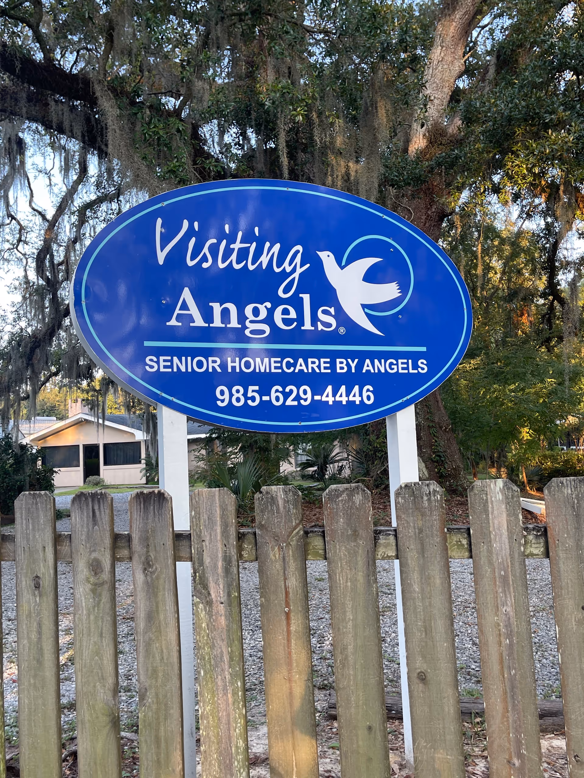 Blue Visiting Angels sign with logo behind a wooden fence and trees.