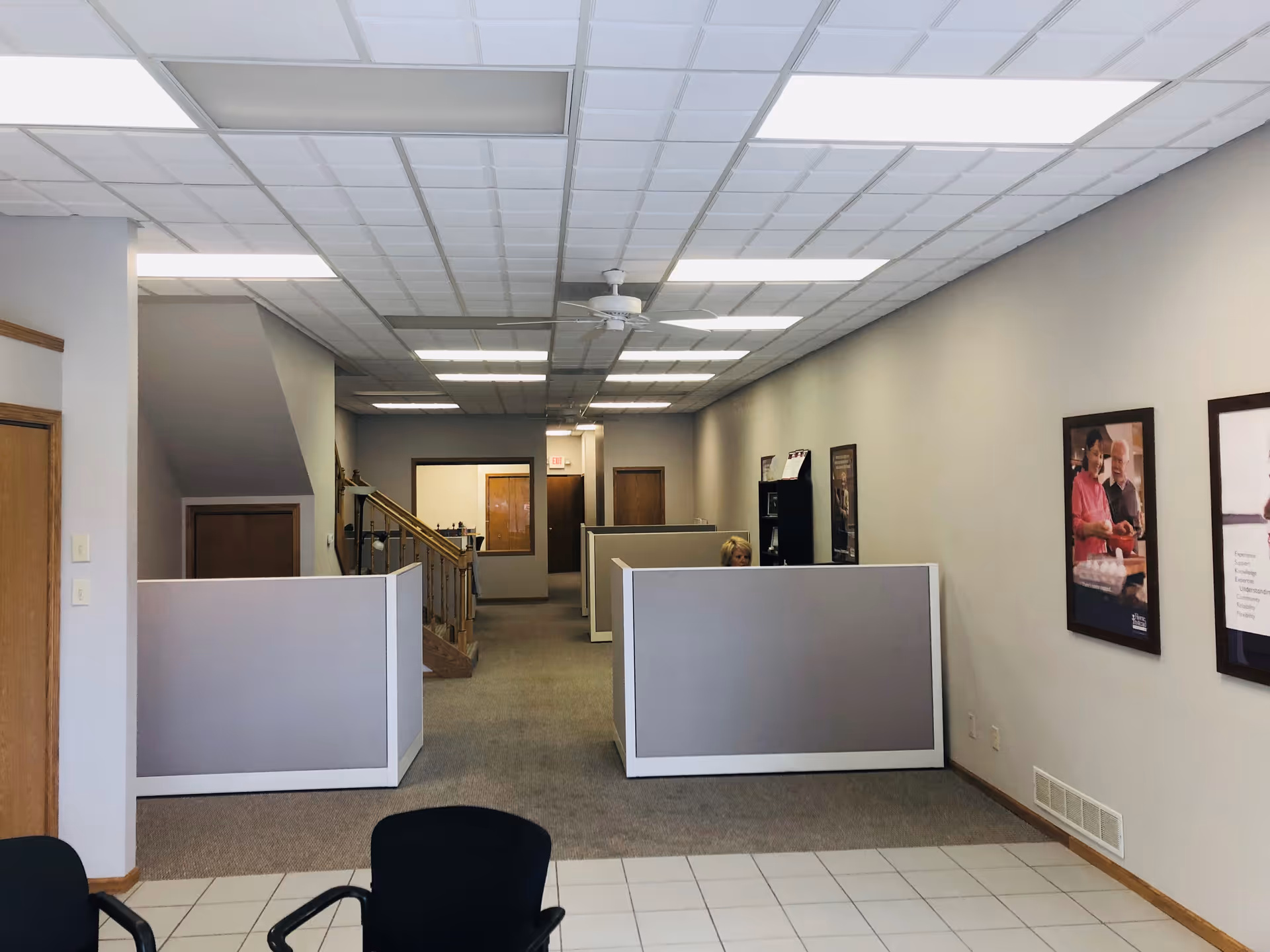 Interior office area with gray cubicles, a staff member, and framed posters along the wall.