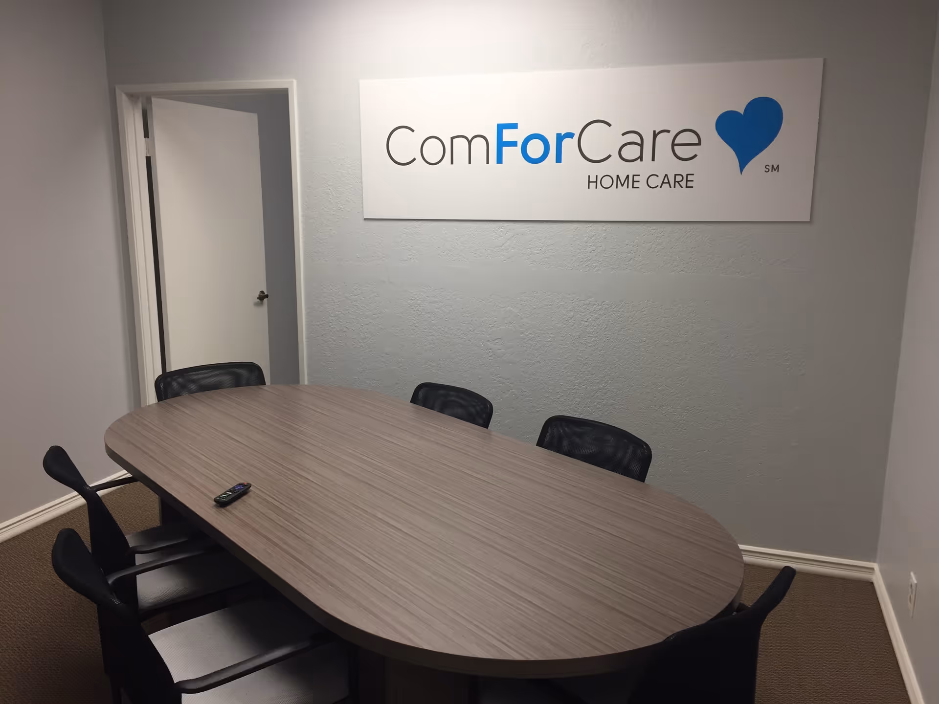 An office conference room with oval table, black chairs, and ComForCare sign on wall.