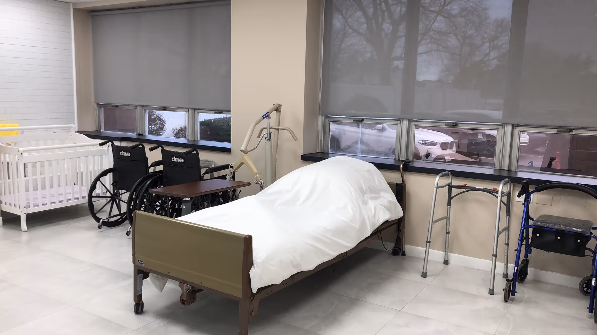 An empty hospital-style patient room with a bed, wheelchairs, walkers, and a crib.
