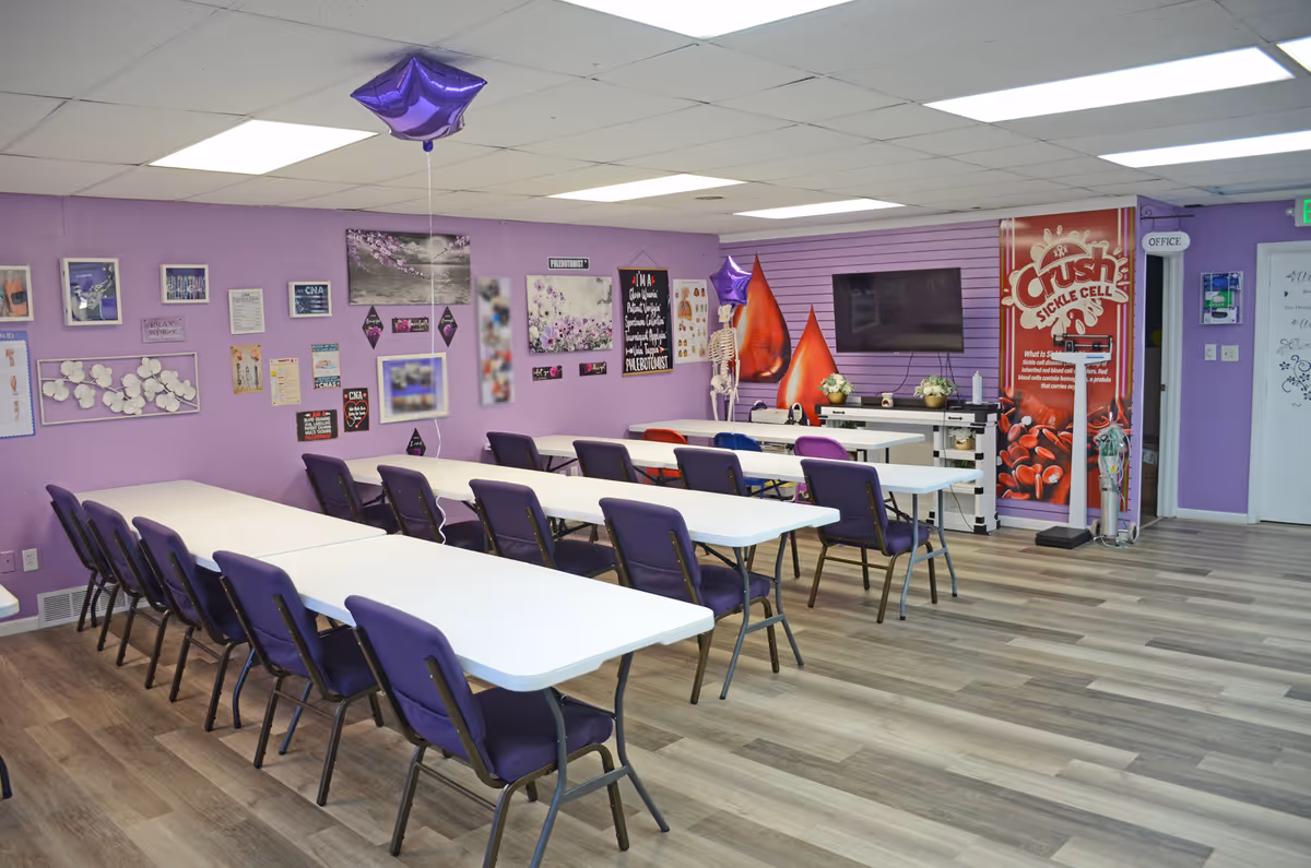 A brightly lit purple-walled dining room with many white tables and purple chairs.