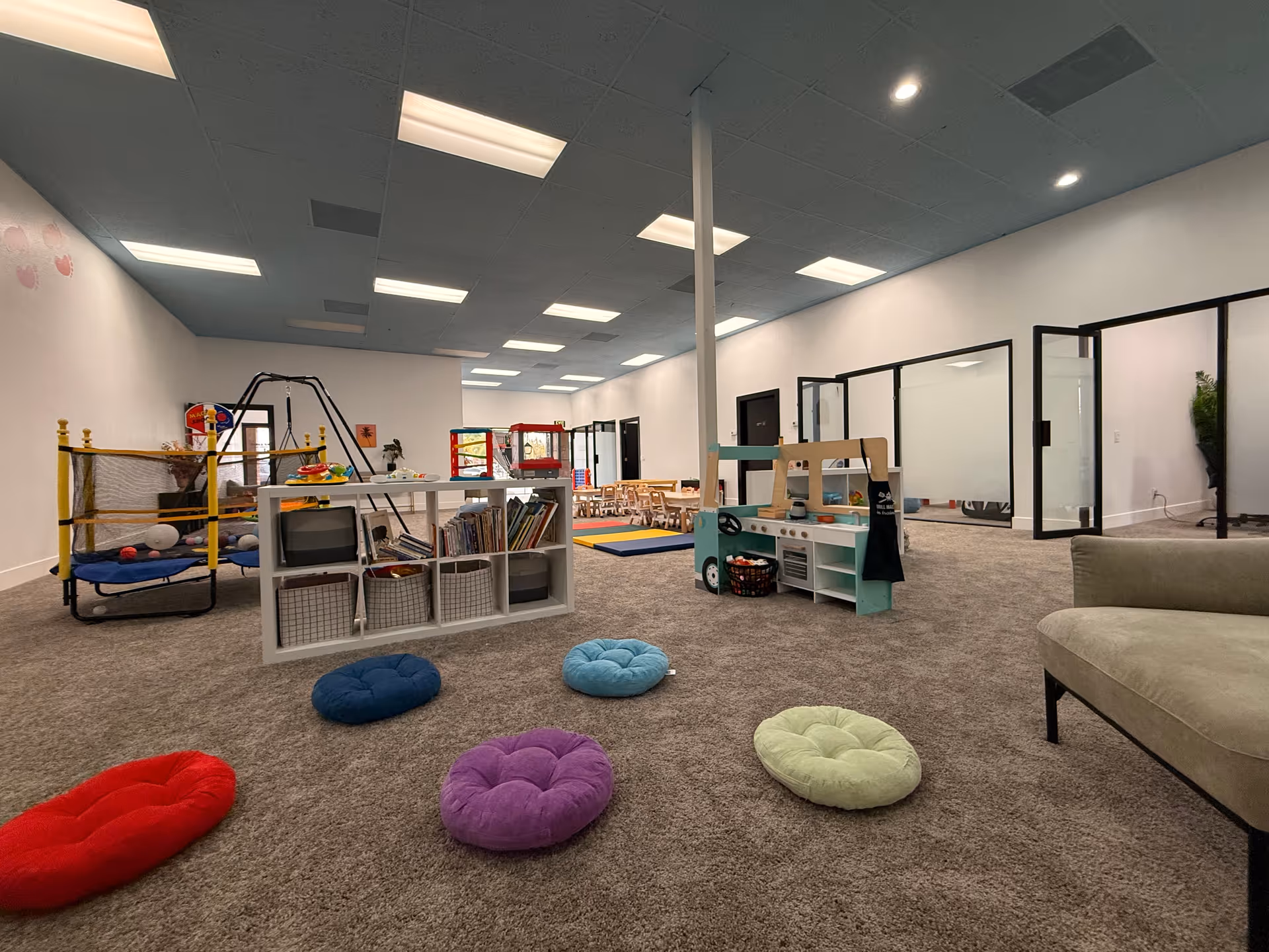 Open indoor play area with shelves, climbing structure, colorful cushions, and glass-walled rooms.
