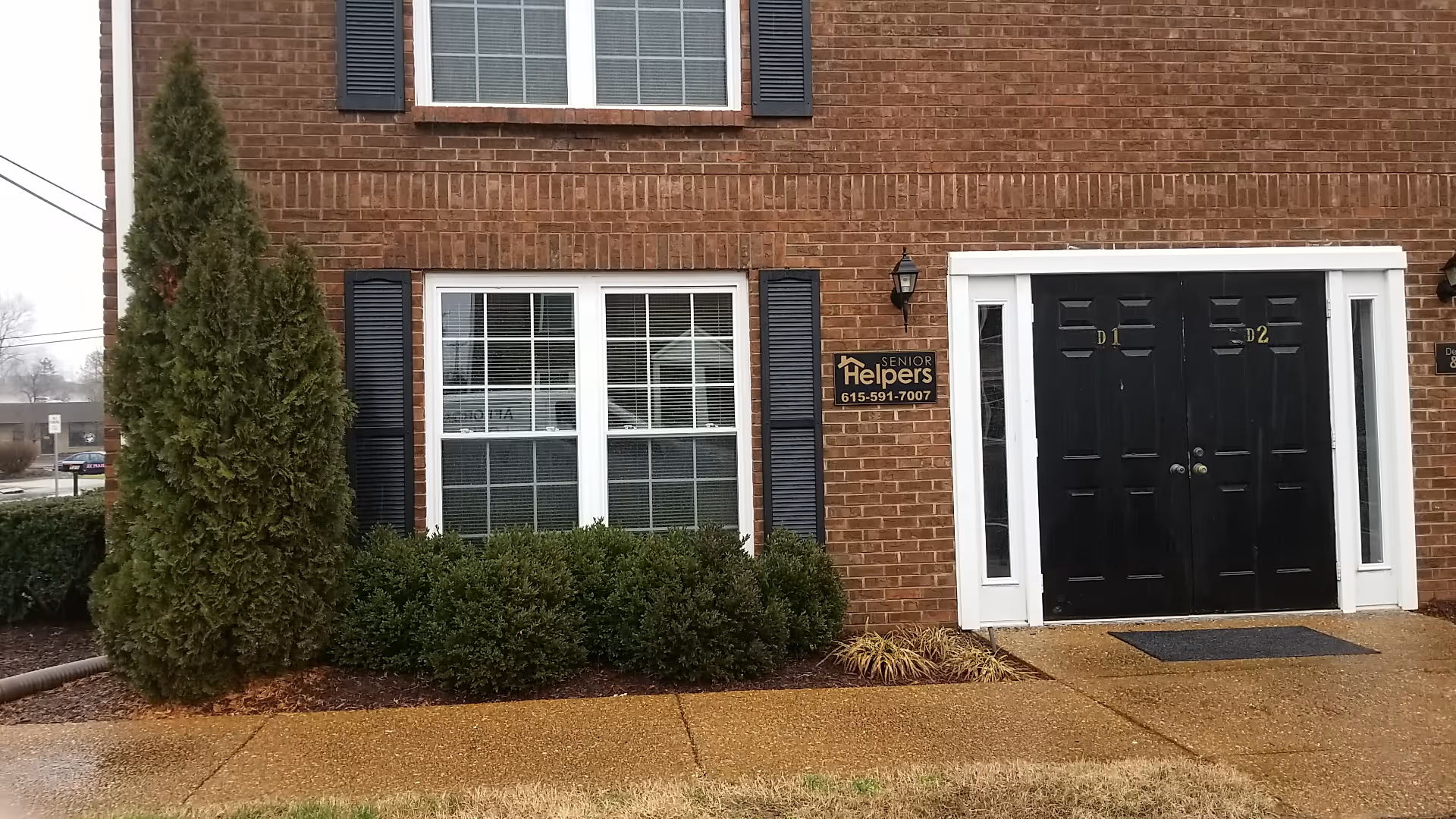 Brick senior living building exterior with dark double doors, windows, shrubs, and a Senior Helpers sign.