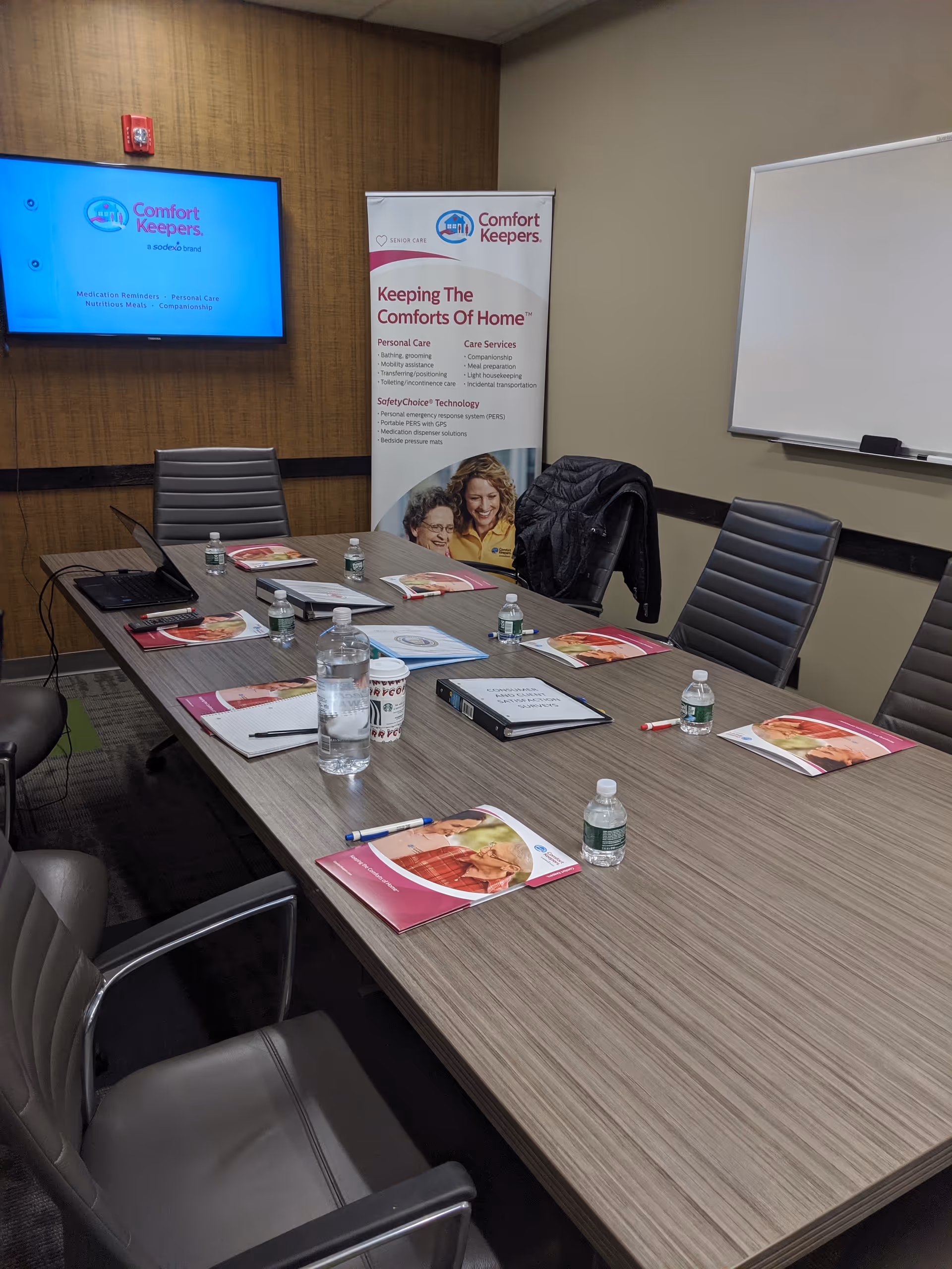 Conference room with long table, Comfort Keepers banners, brochures, water bottles, and a whiteboard.