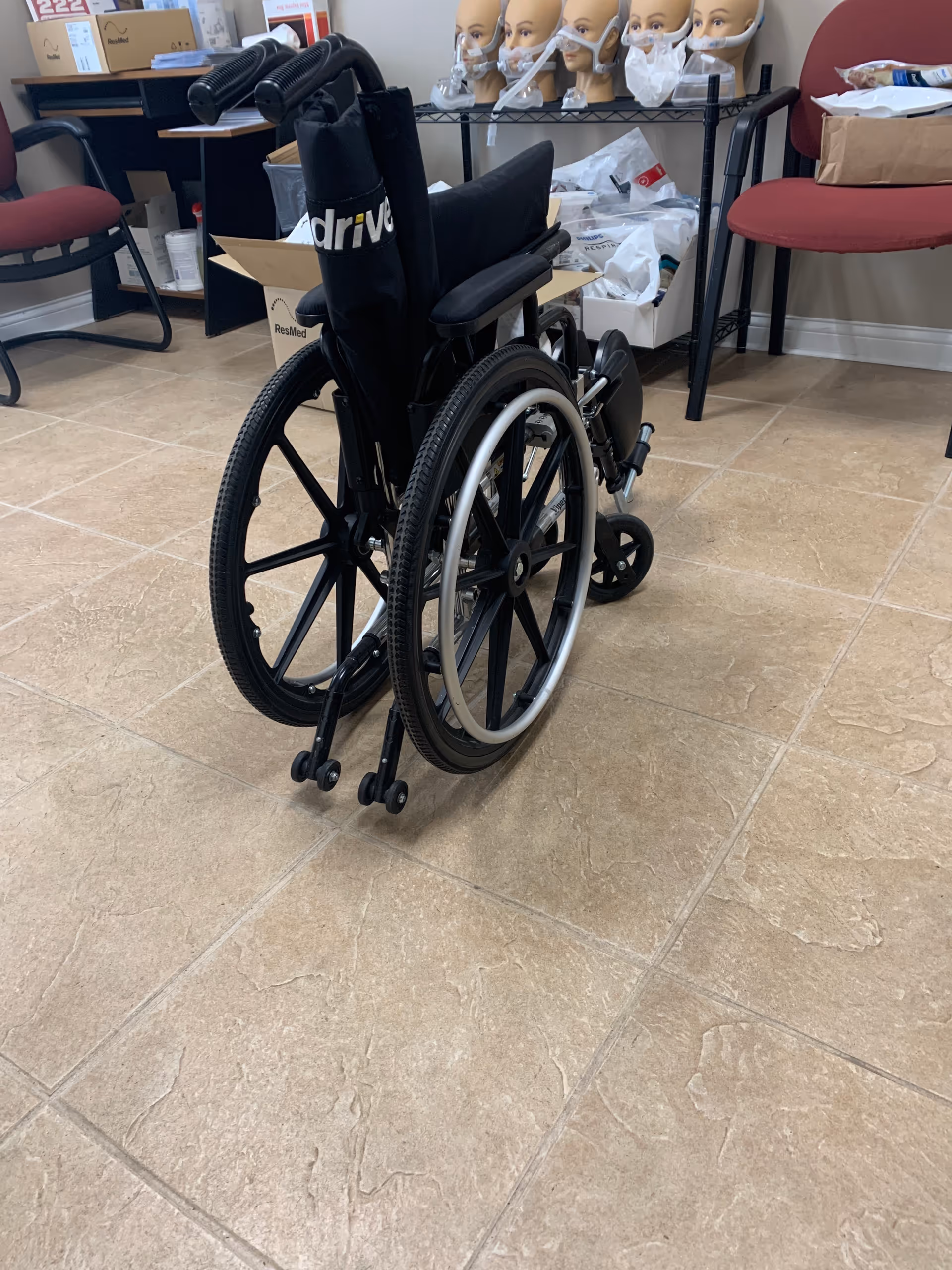 An empty wheelchair sits in a LINCARE facility room with medical mannequins and supplies.